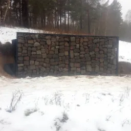Gabionowa ściana oporowa z kamienia w metalowej siatce, zabezpieczająca skarpę pokrytą śniegiem w zimowym krajobrazie.