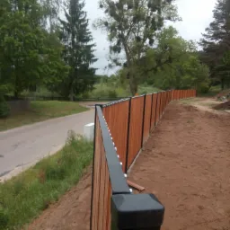 Nowo postawiony drewniany płot z czarnymi elementami wykończeniowymi, biegnący wzdłuż drogi i ziemistego terenu, z drzewami w tle.