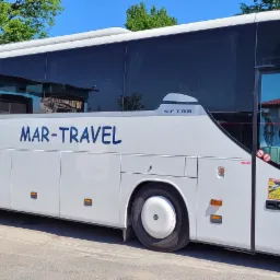 Biały autobus turystyczny marki Setra z niebieskim napisem 'MAR-TRAVEL' zaparkowany przed budynkiem z niebieską fasadą.