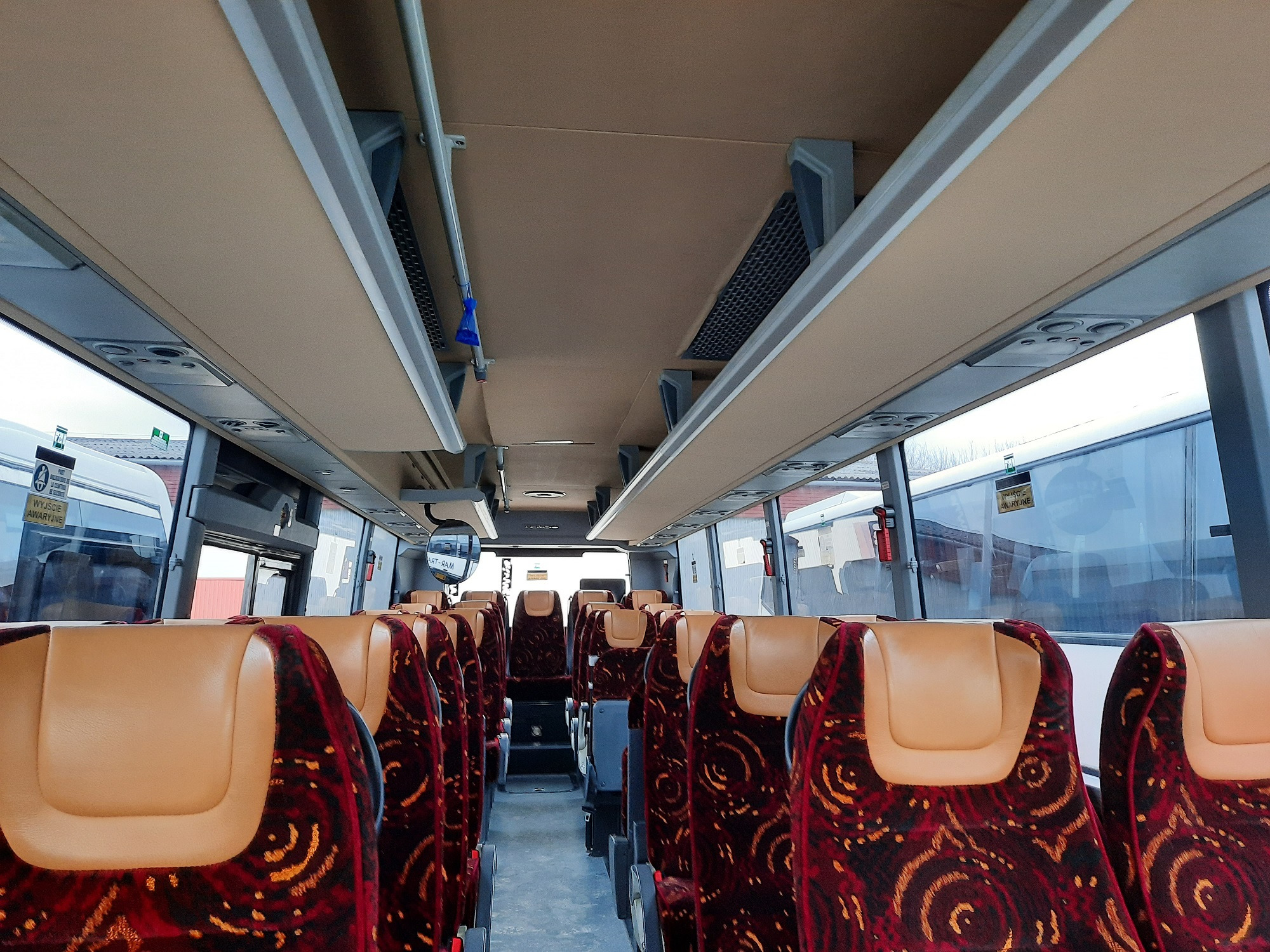 Wnętrze autobusu w Strzelinie z beżowymi zagłówkami i czerwono-złotymi wzorzystymi siedzeniami widziane z perspektywy pasażera, z widocznymi oknami i elementami oświetlenia sufitowego.