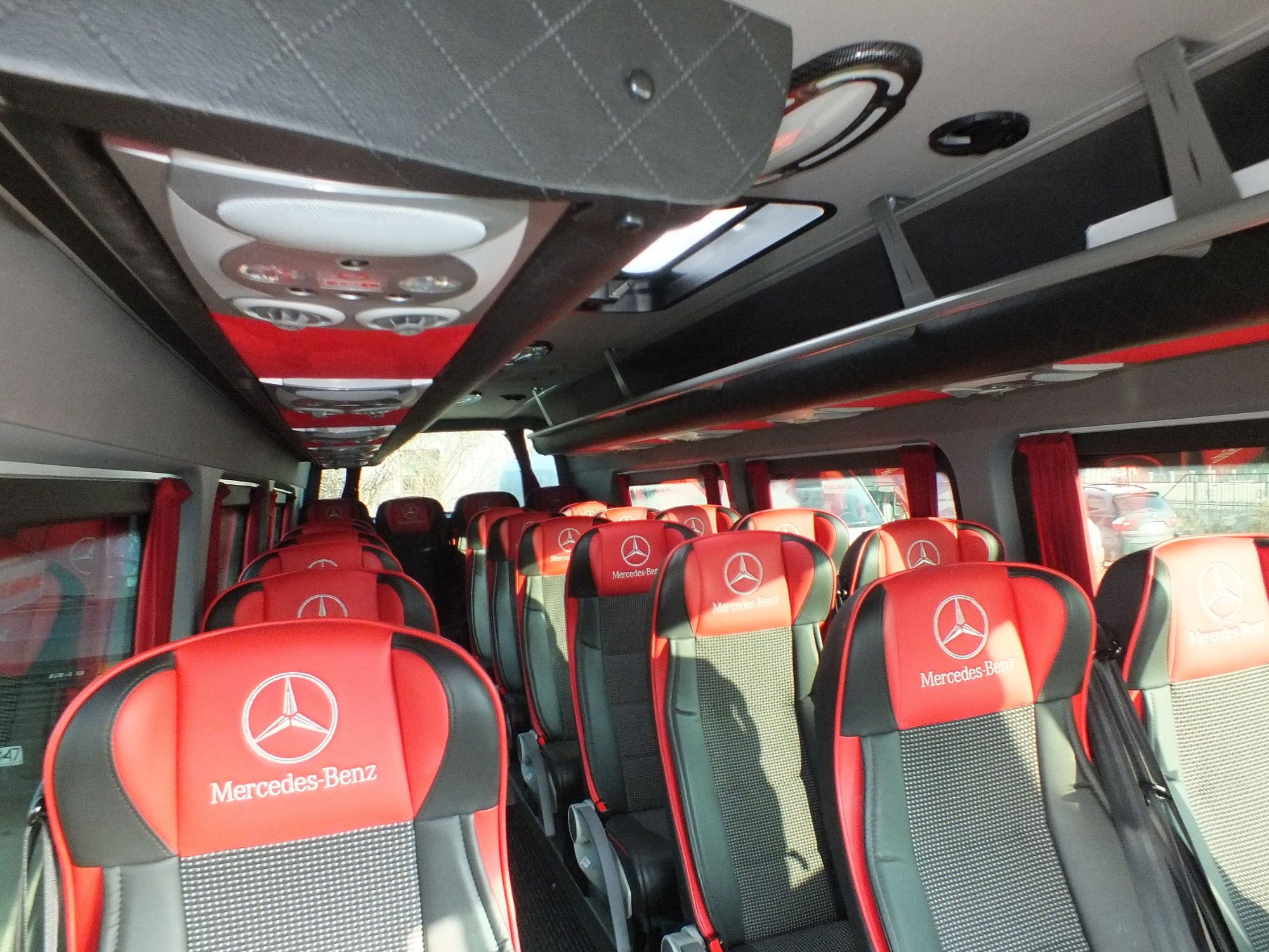 Wnętrze luksusowego busa Mercedes-Benz z czerwono-czarnymi fotelami, widok z perspektywy pasażera w kierunku przodu pojazdu, oświetlenie sufitowe włączone.