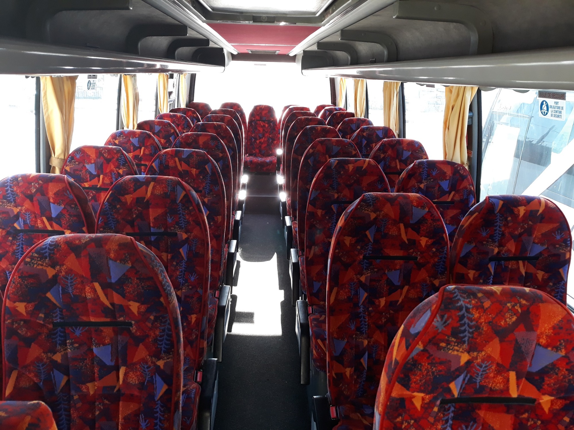 Wnętrze autobusu z rzędami foteli obitych wzorzystą tapicerką w odcieniach czerwieni, pomarańczy i granatu, widok wzdłuż korytarza w kierunku jasnego światła z przodu pojazdu.