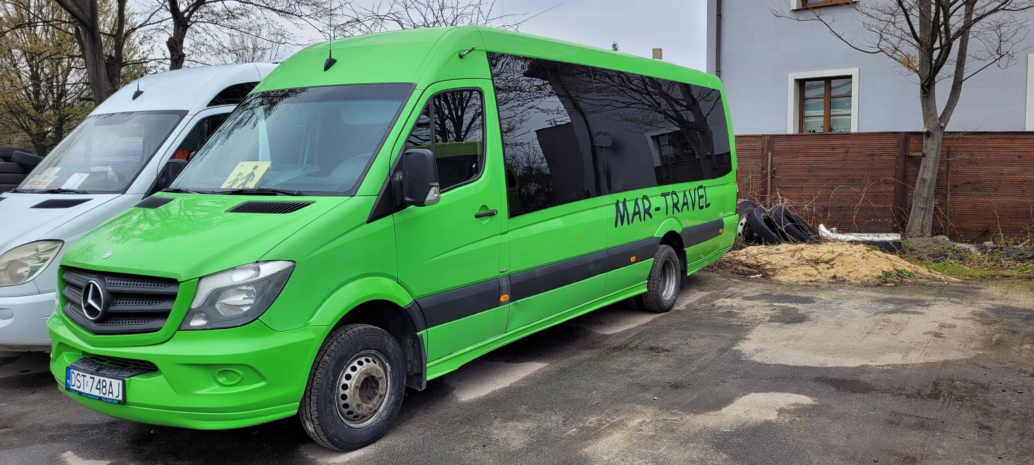 Zielony bus Mercedes-Benz Sprinter z napisem 'MAR-TRAVEL' na boku, zaparkowany na zewnątrz, widoczny z boku, obok stoi biały bus.