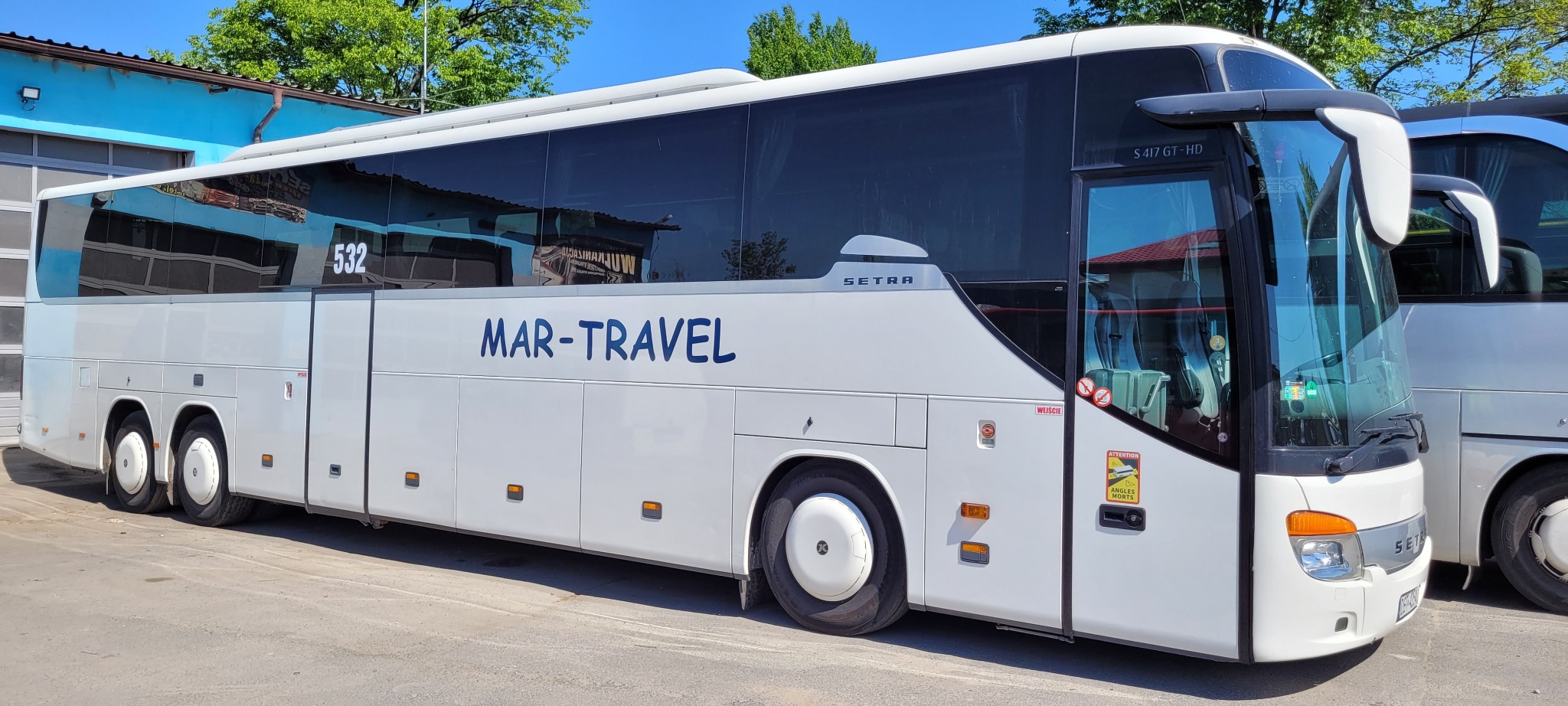 Biały autobus turystyczny marki Setra z niebieskim napisem 'MAR-TRAVEL' zaparkowany przed budynkiem z niebieską fasadą.