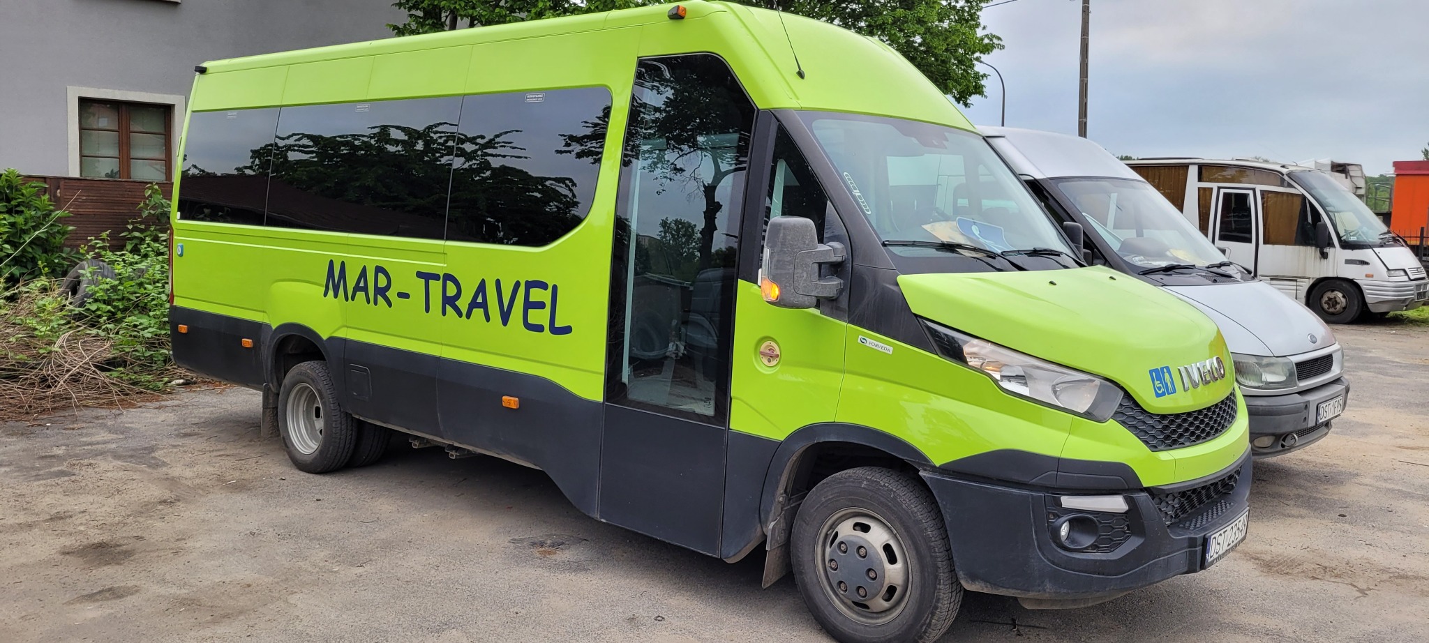 Jasnozielony bus z napisem MAR-TRAVEL, zaparkowany obok innych busów na placu.