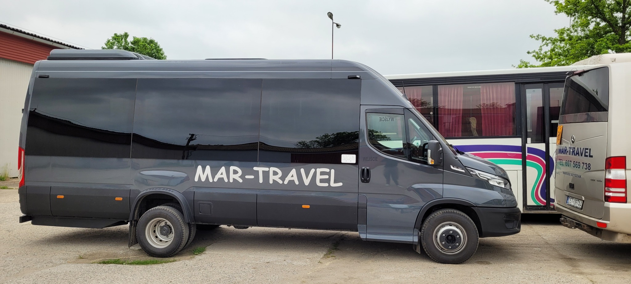 Szary bus marki Iveco z białym napisem 'MAR-TRAVEL' na boku, stojący na parkingu obok innych autobusów, widok z boku w pochmurny dzień.