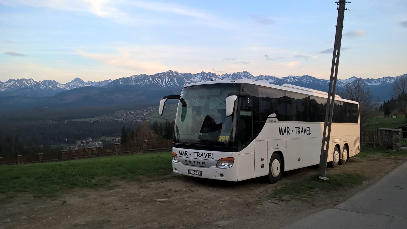 Biały autokar Setra MAR-TRAVEL na tle górskiego krajobrazu z widocznymi ośnieżonymi szczytami i zabudowaniami w dolinie.