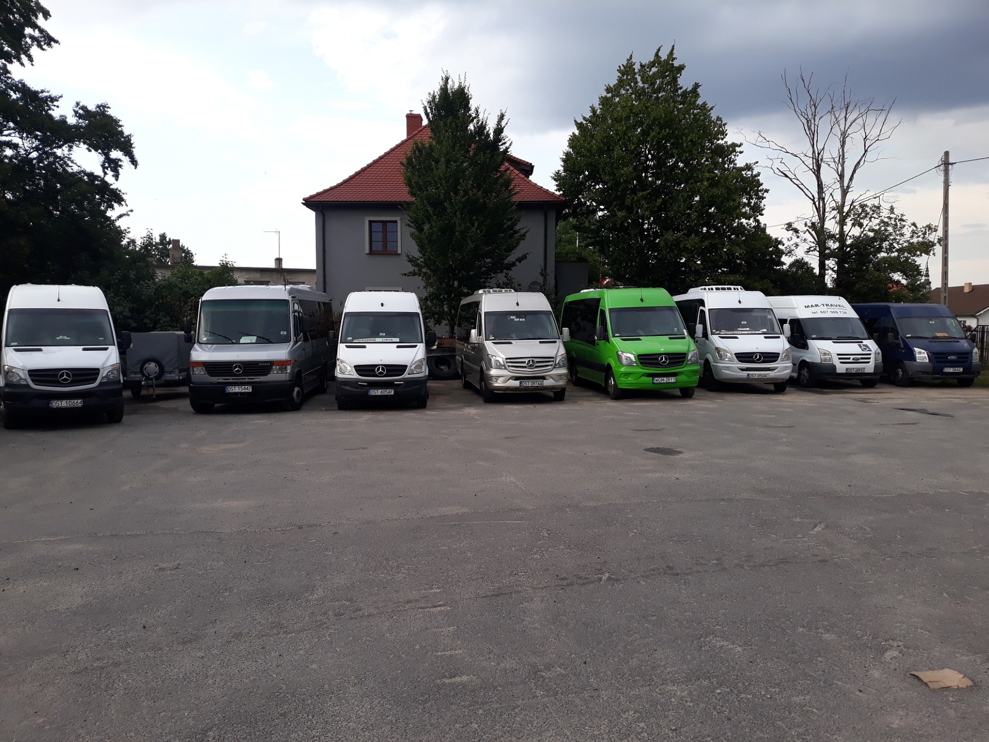 Flota różnokolorowych busów Mercedes-Benz zaparkowana na placu przed budynkiem z czerwoną dachówką w pochmurny dzień. Widoczne numery rejestracyjne i logo firmy MAR-TRAVEL na jednym z busów.