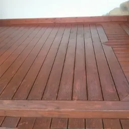 Ciemnobrązowa drewniana podłoga tarasu z dekoracyjnym wzorem w kształcie rombu, otoczona kamienną ścianą i białą ścianą budynku.