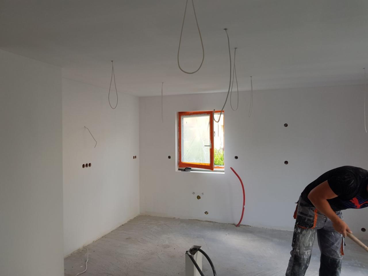 Wnętrze w trakcie malowania na biało, widoczne wystające kable, okno z pomarańczową ramą i pracownik wygładzający podłogę.