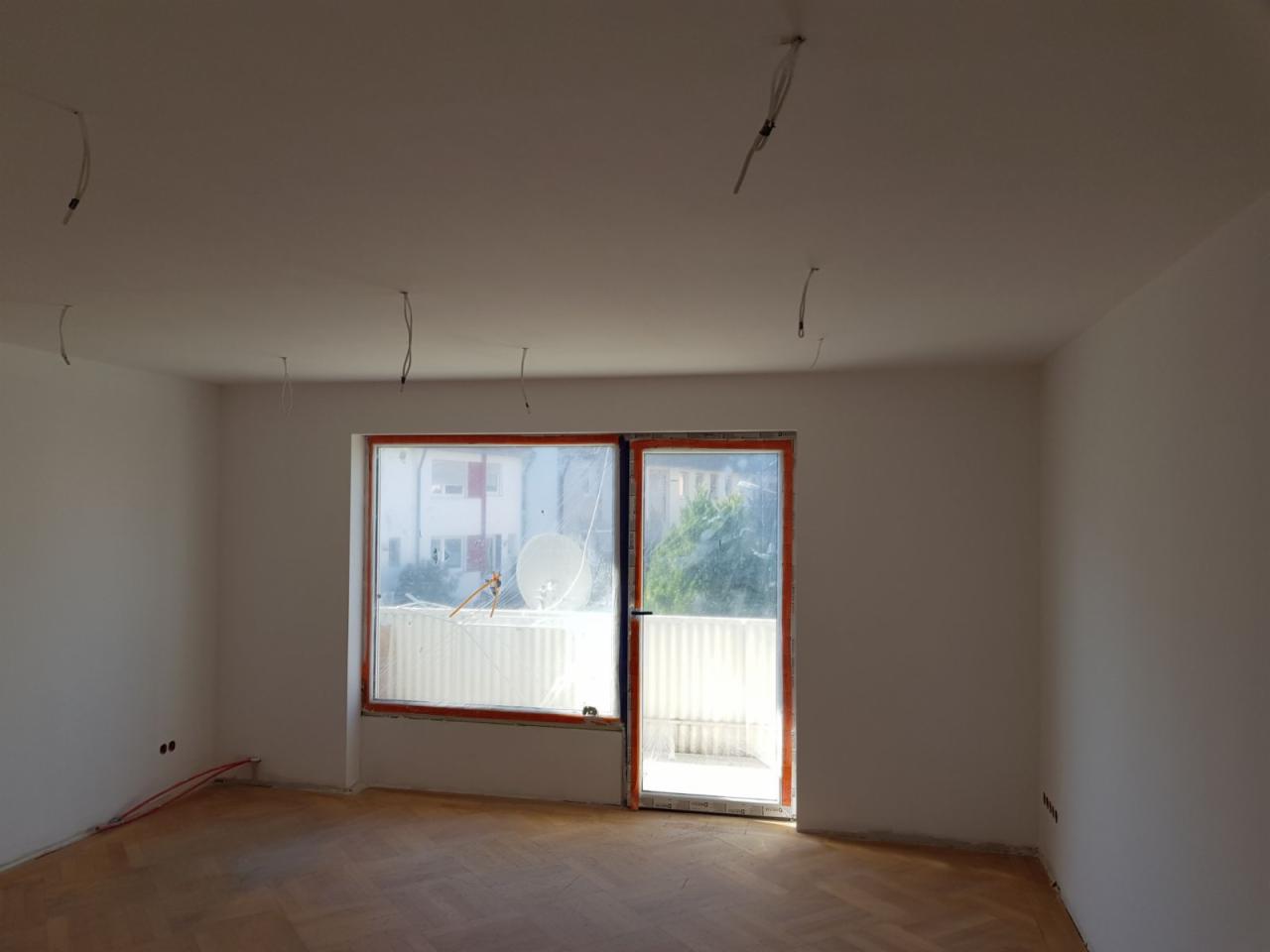 Wnętrze pokoju w trakcie prac wykończeniowych: świeżo pomalowane ściany na biało, parkiet ułożony w jodełkę, okno balkonowe z pomarańczową ramą zabezpieczone folią malarską, wystające przewody...