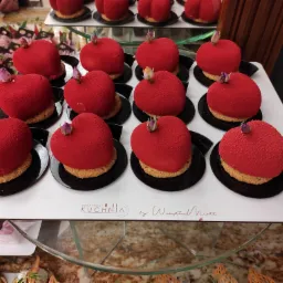 Rząd czerwonych deserów w kształcie serca z teksturą przypominającą aksamit, umieszczonych na kruchym spodzie i czarnych talerzykach, udekorowanych drobnymi pąkami kwiatów, prezentowanych na białej...