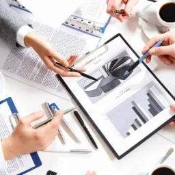 Zespół analizuje wykresy na białym stole, widoczne dłonie wskazujące na dane, filiżanki kawy i dokumenty w tle.