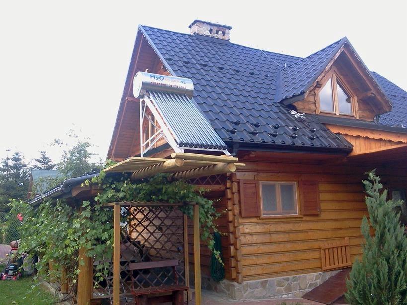 Kolektor słoneczny cylindryczny zamontowany na dachu drewnianego domu z ciemną dachówką, widok z poziomu ogrodu z altaną.