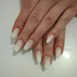 Dłonie z białymi, ombre paznokciami w kształcie trumienki, ozdobionymi rzędem drobnych, błyszczących cyrkonii przy nasadzie.
