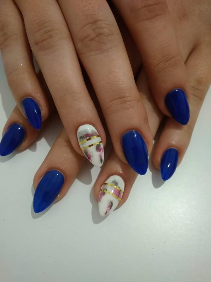 Dłonie z pomalowanymi paznokciami w odcieniach granatu i bieli, dwa paznokcie z abstrakcyjnym wzorem i złotymi paskami.