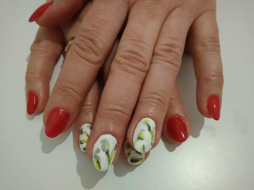 Dłonie z pomalowanymi paznokciami: czerwony lakier na niektórych paznokciach i biały lakier z abstrakcyjnym wzorem w odcieniach szarości i żółci na pozostałych, owalny kształt paznokci.