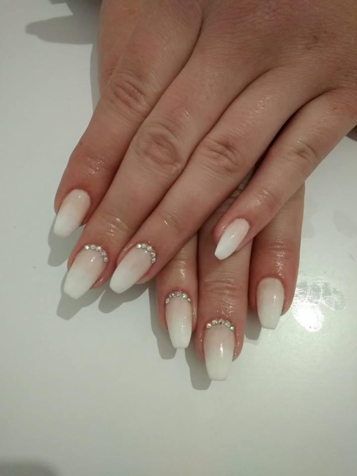 Dłonie z białymi, ombre paznokciami w kształcie trumienki, ozdobionymi rzędem drobnych, błyszczących cyrkonii przy nasadzie.