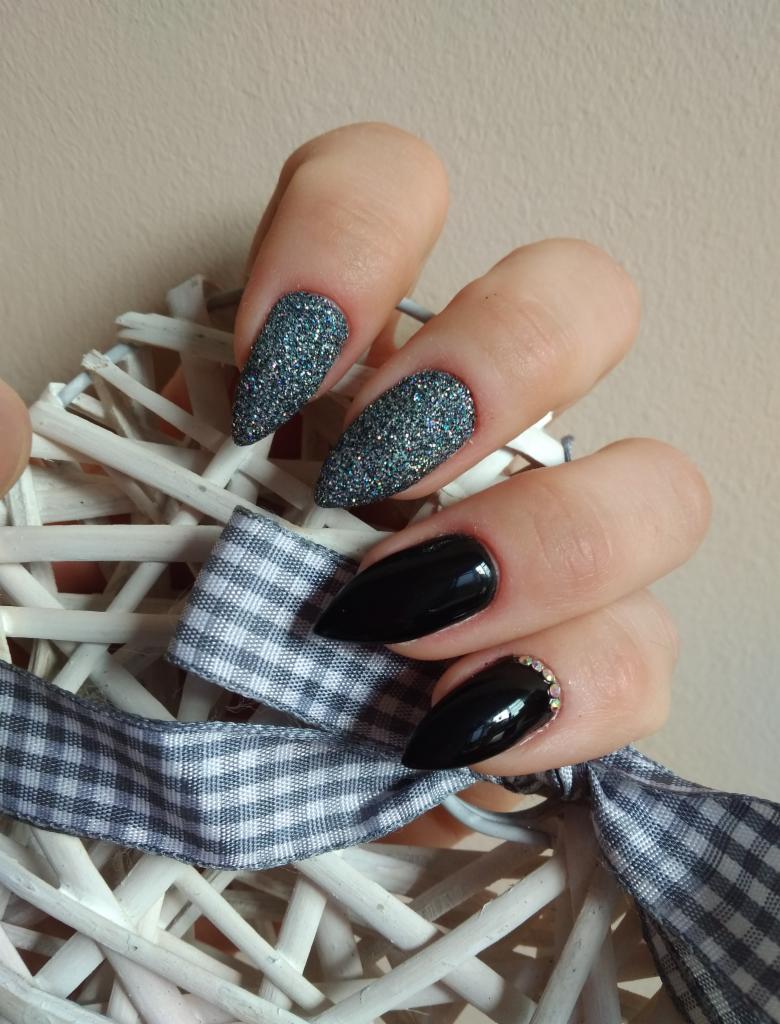 Dłoń z pomalowanymi paznokciami w kształcie szpiców: dwa pokryte brokatem w odcieniach szarości, jeden czarny z drobnymi kryształkami u nasady, dłoń ułożona na białym wiklinowym sercu z szarą...