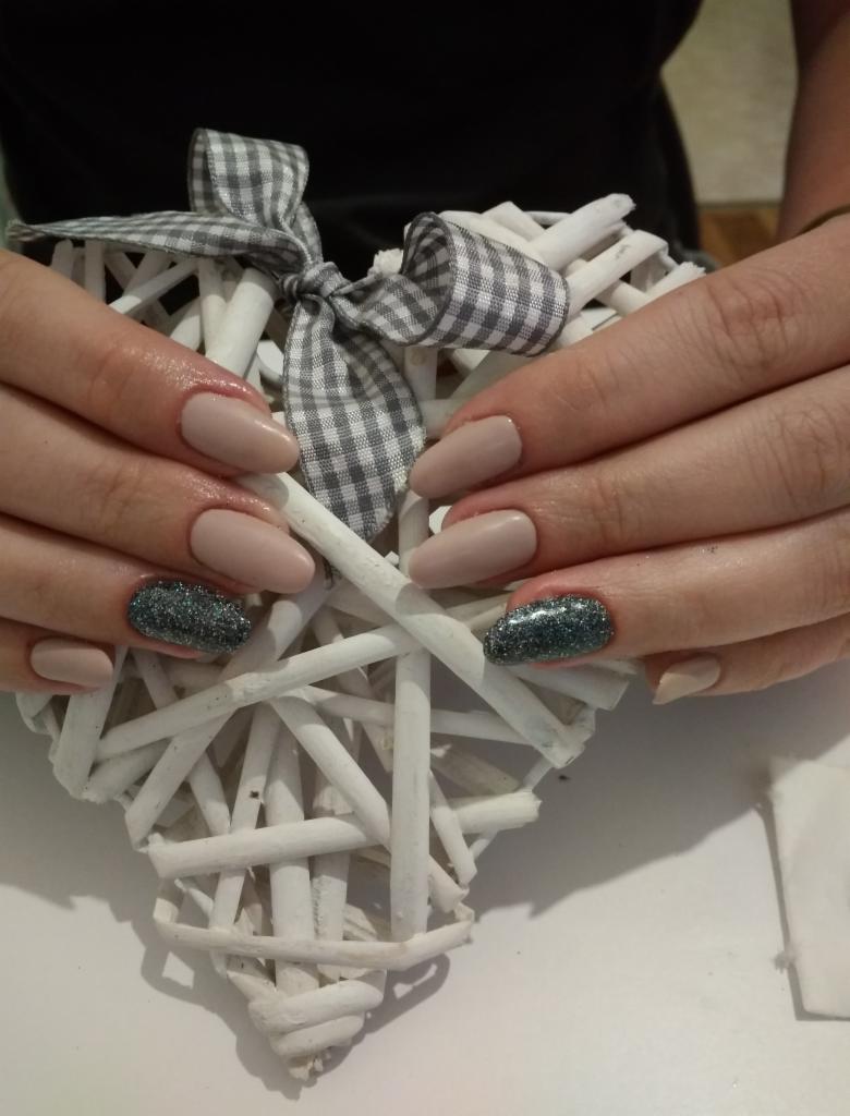 Dłonie z beżowymi paznokciami, jeden paznokieć na każdej dłoni z brokatowym zdobieniem, na tle białego serca z wikliny z szarą kokardą w kratkę.