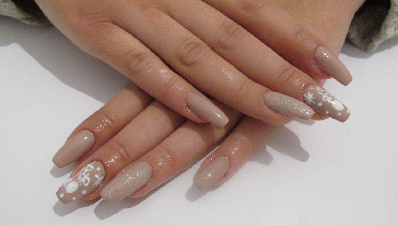 Dłonie z beżowymi paznokciami w kształcie migdała, na jednym paznokciu wzór śnieżynki i bałwana, na innym brokatowy lakier.