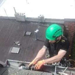 Pracownik w zielonym kasku, zabezpieczony linami, wykonuje prace na dachu, prawdopodobnie naprawiając rynnę, widok z góry na pochylony dach i fragment budynku z cegły.
