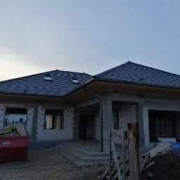 Surowy dom jednorodzinny w trakcie budowy z ciemnym, nowym dachem z dachówką ceramiczną, widoczne okna dachowe i częściowo wykonana elewacja z pustaków. Na pierwszym planie czerwony kontener...