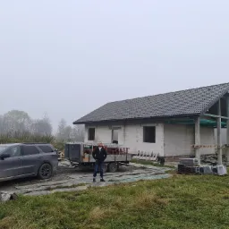 Niedokończony dom jednorodzinny z szarej cegły z zamontowanym dachem z ciemnej dachówki, na posesji stoi szary SUV i przyczepa, a mężczyzna w czarnej kurtce stoi przed przyczepą na tle mglistego...