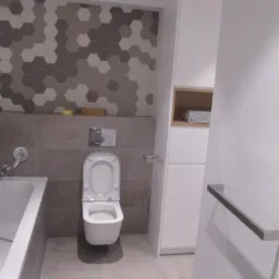 Nowoczesna łazienka z mozaiką heksagonalną w odcieniach szarości, wisząca toaleta z otwartą klapą, biała zabudowa meblowa i szara podłoga z płytek.