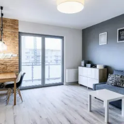 Jasne wnętrze mieszkania z drewnianą podłogą, ceglaną ścianą z neonowym napisem 'happy', szarą ścianą z trzema obrazkami i szarą sofą.