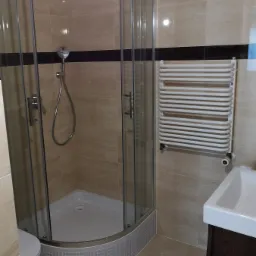 Wykończona łazienka z kabiną prysznicową z zaokrąglonymi szybami, grzejnikiem drabinkowym, umywalką z szafką oraz beżowymi płytkami ściennymi z ciemnobrązowym pasem dekoracyjnym.