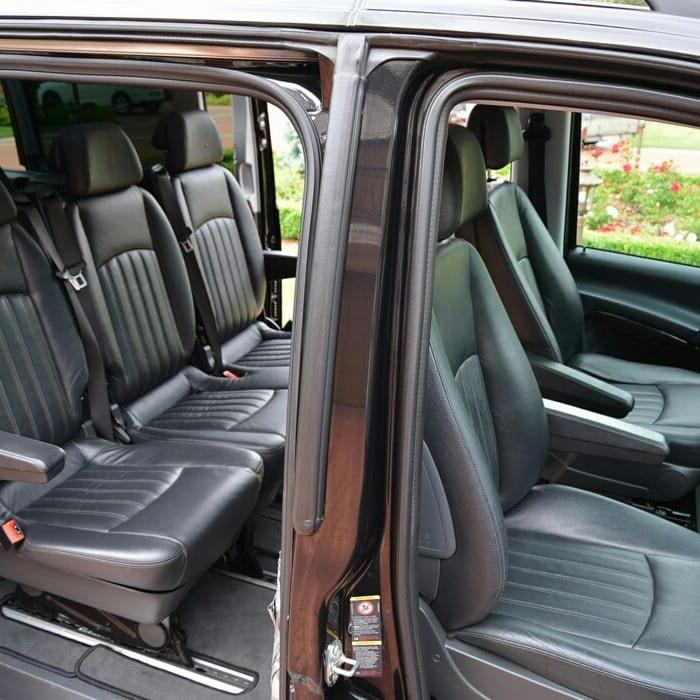 Wnętrze czarnego busa z luksusowymi skórzanymi siedzeniami i otwartymi drzwiami, widok na zadbany ogród w tle.