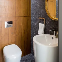 Nowoczesna łazienka z drewnianymi panelami, umywalką na postumencie i okrągłym lustrem w złotej ramie, z widocznym fragmentem toalety podwieszanej.