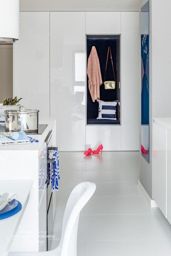 Biała kuchnia z zabudową szaf wnękowych, wnęka z wieszakiem na ubrania, torebką i poduszką w stylu marynistycznym, różowe szpilki na podłodze.