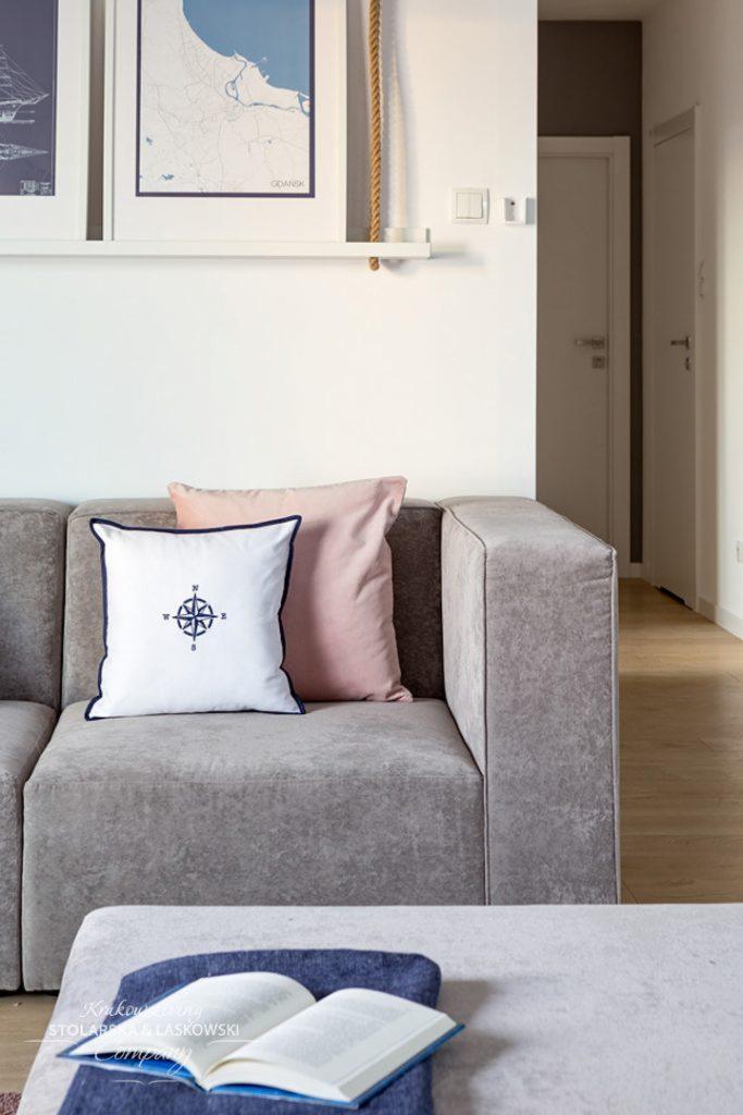 Szary modułowy narożnik z poduszkami w pastelowych kolorach, biała poduszka z motywem kompasu, otwarta książka na pufie, na ścianie mapa Gdańska w ramie i lina dekoracyjna.
