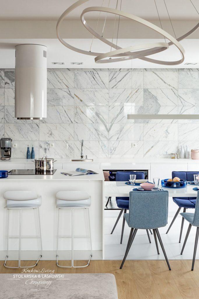 Nowoczesna kuchnia z białymi meblami, marmurową ścianą i okrągłym, designerskim oświetleniem sufitowym; widoczne wysokie, białe hokery i stół z niebieskimi krzesłami.