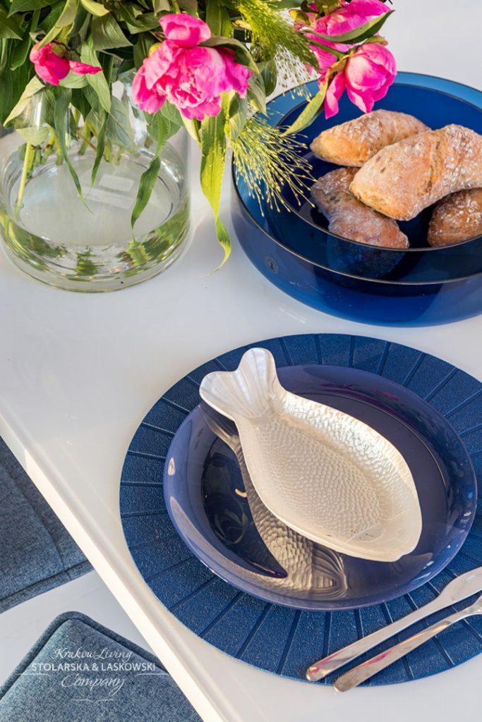 Dekoracyjny stół z białym blatem, niebieską zastawą, srebrnym półmiskiem w kształcie ryby, pieczywem w niebieskiej misie i wazonem z różowymi piwoniami.