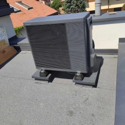 JAMRÓZ Nowoczesne Technologie - Szara pompa ciepła Panasonic Aquarea A, zamontowana na płaskim dachu, z widocznymi elementami mocującymi i fragmentami dachu budynku w tle.