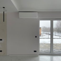 JAMRÓZ Nowoczesne Technologie - Biały klimatyzator na ścianie w jasnym wnętrzu, widoczne okno z widokiem na zaśnieżony ogród. Minimalistyczny design, nowoczesne wykończenie, widoczne gniazdka elektryczne.