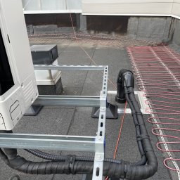 JAMRÓZ Nowoczesne Technologie - Instalacja pompy ciepła na dachu budynku z widocznymi rurami i elementami montażowymi, z bliska. Widoczne przewody grzewcze na dachu.