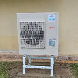 Zewnętrzna jednostka klimatyzacji Atlantic na metalowym stelażu, zamontowana przy budynku z beżową elewacją i widocznym logo firmy Jamróz. Widoczna trawa i kostka brukowa.