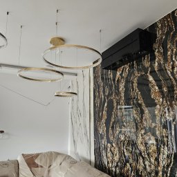 JAMRÓZ Nowoczesne Technologie - Nowoczesny klimatyzator ścienny w czarnej obudowie, zamontowany na ścianie wykończonej dekoracyjną płytą imitującą marmur. Widoczne okrągłe lampy w folii ochronnej.