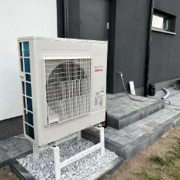 Zewnętrzna jednostka klimatyzacji Atlantic DC Inverter na białym stelażu, z szarymi kamykami pod spodem, na tle szarego budynku z czarnymi drzwiami.