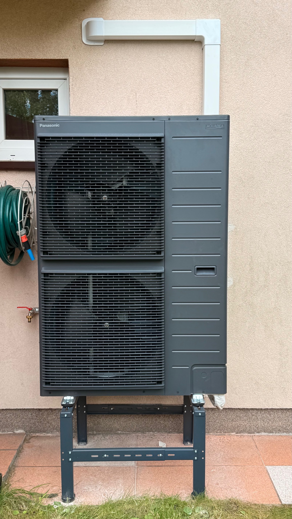 Szara pompa ciepła Panasonic Aquarea zamontowana na metalowym stelażu przy beżowej ścianie budynku. Widoczny fragment okna i zwinięty wąż ogrodowy.