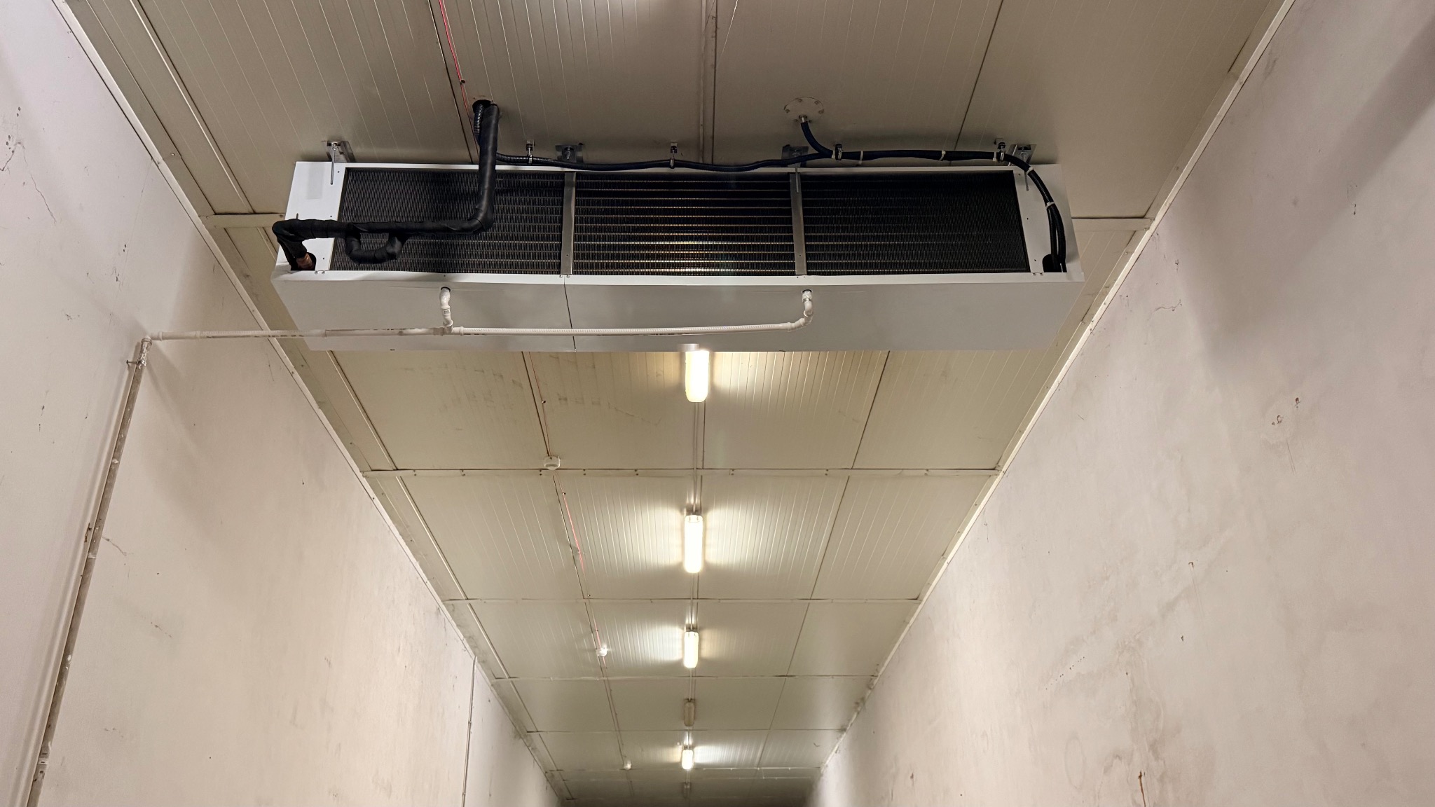 Biała jednostka klimatyzacji przemysłowej zamontowana pod sufitem w hali, z widocznymi rurami i przewodami, oświetlona szeregiem lamp.
