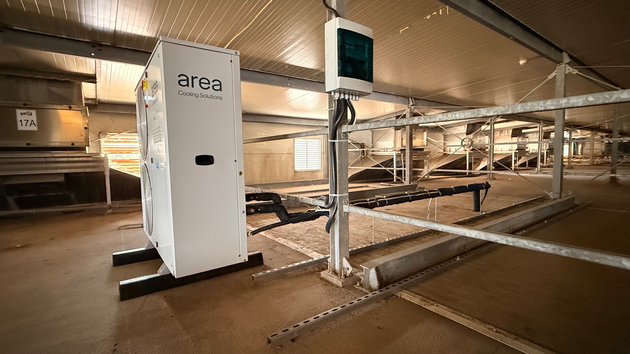 Biała jednostka klimatyzacji przemysłowej 'Area Cooling Solutions' w hali z metalową konstrukcją. Widoczne przewody i skrzynka elektryczna. Oświetlenie górne.