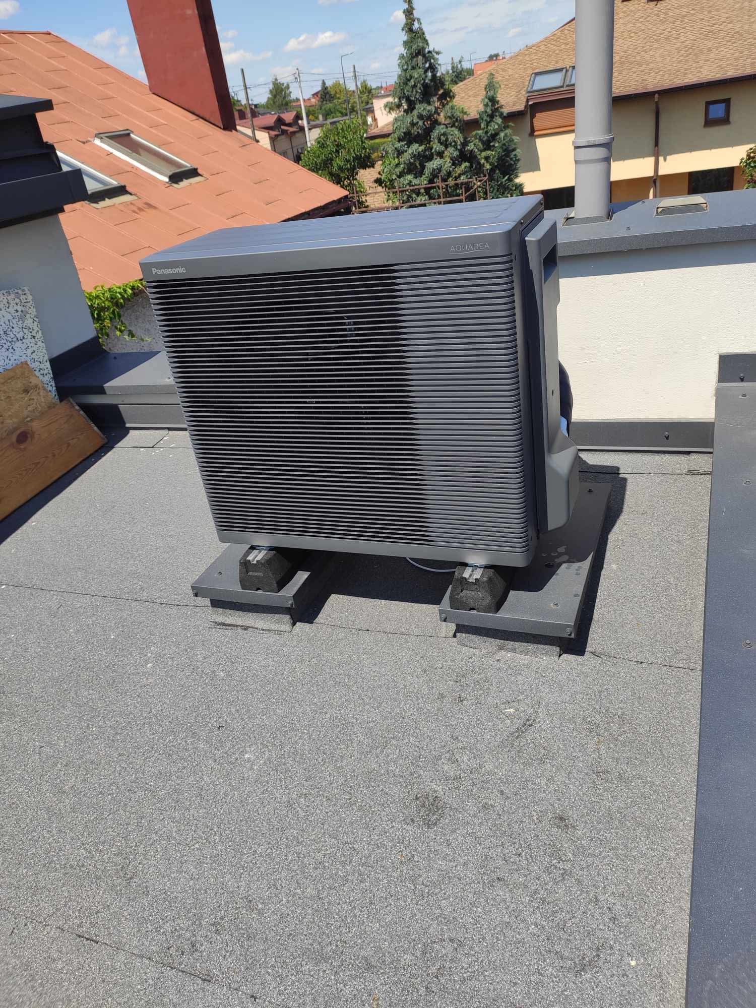 Szara pompa ciepła Panasonic Aquarea A, zamontowana na płaskim dachu, z widocznymi elementami mocującymi i fragmentami dachu budynku w tle.
