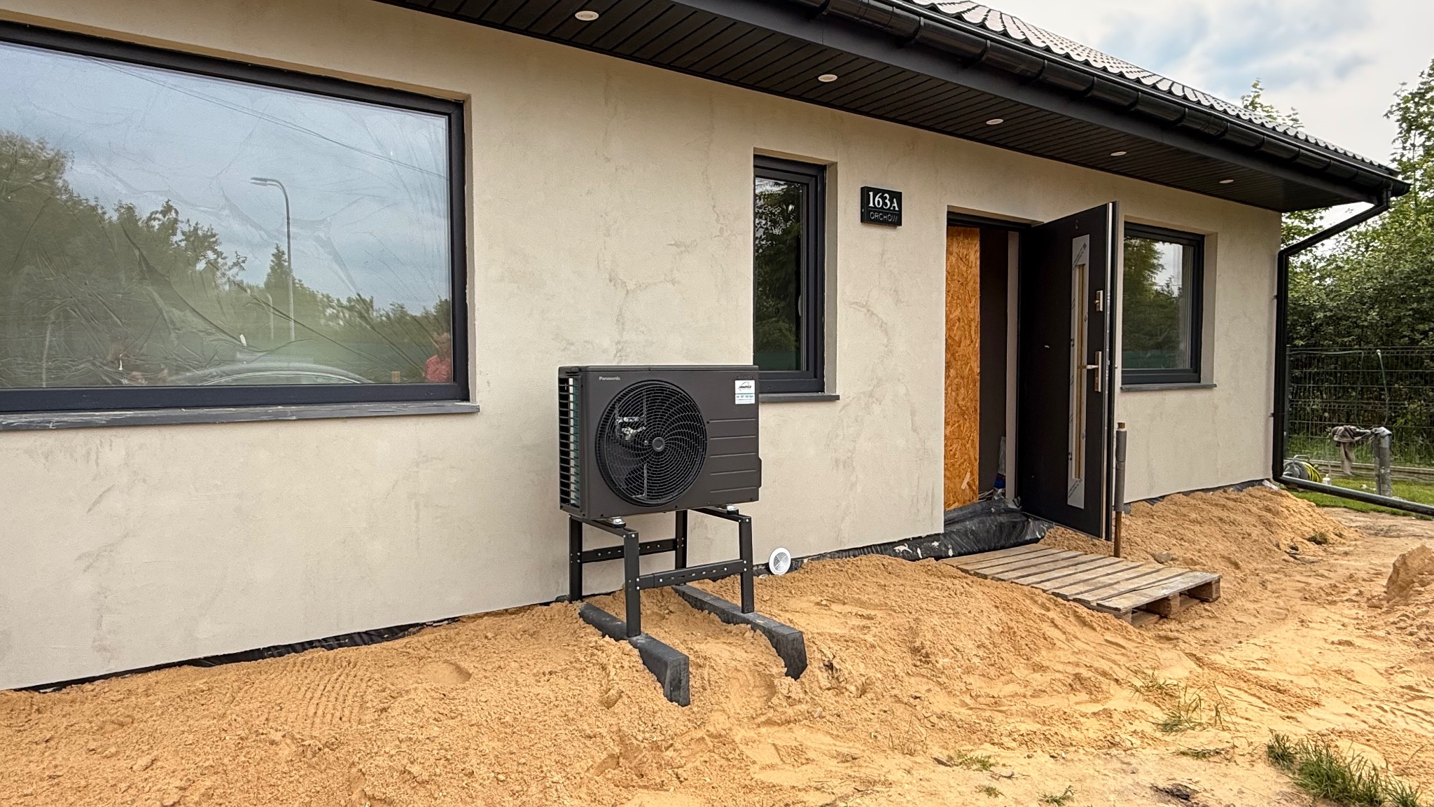 Pompa ciepła Panasonic zamontowana na zewnątrz budynku z beżową elewacją, widoczna tabliczka z numerem domu 163A. Teren wokół w trakcie prac ziemnych.