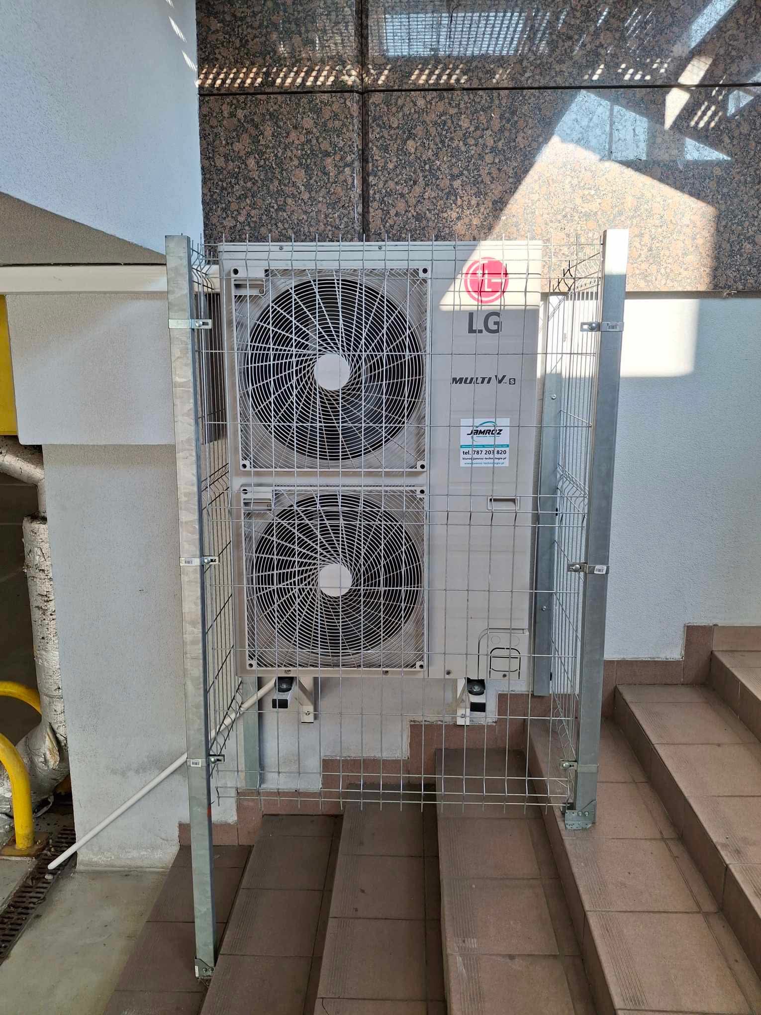 Zewnętrzna jednostka klimatyzacji LG Multi V zabezpieczona metalową klatką na schodach, z widoczną naklejką z danymi kontaktowymi firmy JAMROZ.