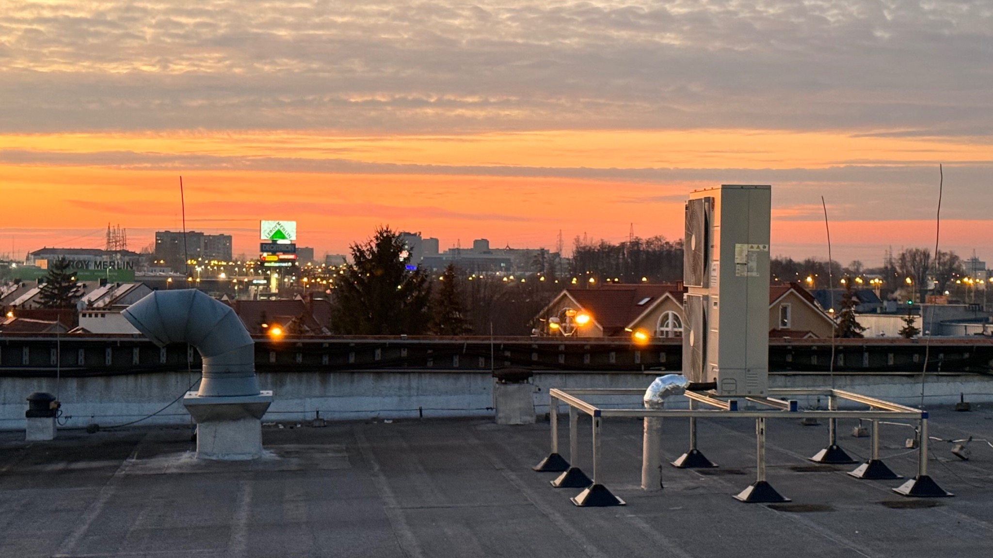 Instalacja klimatyzacji na dachu budynku z widokiem na miasto o zachodzie słońca. Widoczne elementy wentylacyjne i jednostki zewnętrzne klimatyzacji.